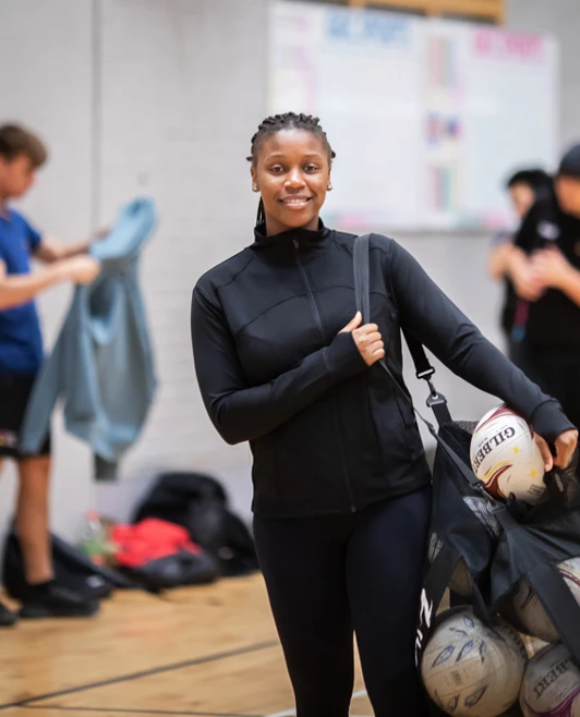 A student holding a mesh bag filled with netballs, smiling at the camera in a sports hall. A student holding a mesh bag filled with netballs, smiling at the camera in a sports hall.